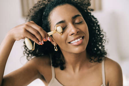 Close up young mixed race woman using jade stone facial roller and smile. Natural self skin care. Beauty routineの写真素材