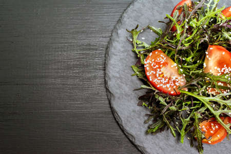 Fresh summer salad with tomatoes, fresh arugula leavesand sesame seeds on black plate over dark wooden background.の写真素材