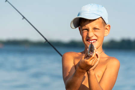 Happy smiling boy holding taken freshwater fish in hands against rod and river backgroundの写真素材