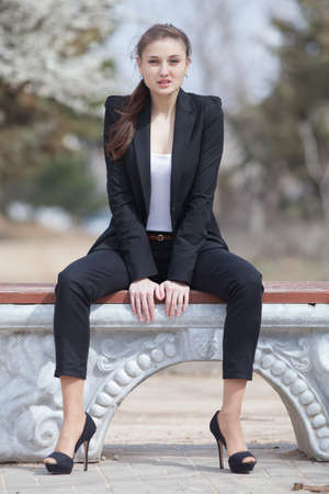 Brunette on stiletto heels outdoors  Young woman in black suit sitting on bench in park looking at cameraの写真素材