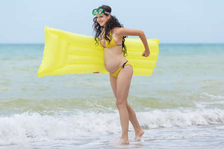 Expectant mother with pool raft and diving mask playing at the sea. Pregnant woman in yellow bikini playing on the beachの写真素材