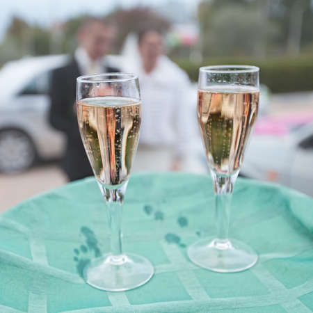 Two wine glasses with champagne on green tablecloth on background of unrecognizable newlywedsの写真素材
