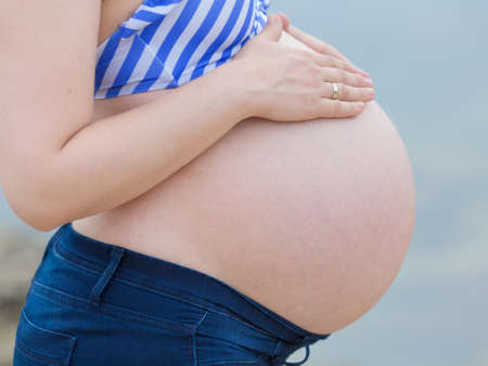 Torso of expectant mother. Pregnant woman in jeans and striped bikini top posing with hands on her naked bellyの写真素材