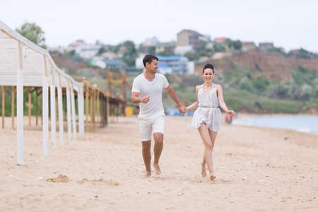 Barefoot couple on sand seashore in cloudy day. Attractive couple in white running holding hands along sand beachの写真素材