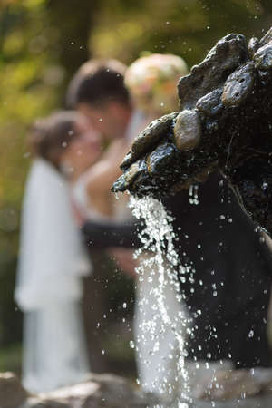 Just married in the park. Newly wedded kissing behind fountain. Focus on foregroundの写真素材