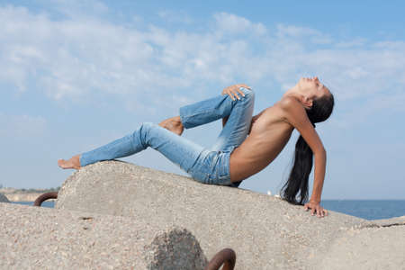 Very slim girl posing on groyne against the sea. Topless young woman in jeans posing on concrete constructionの写真素材