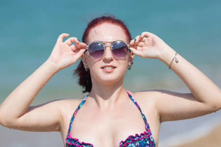 Portrait of red haired girl. Ginger young woman in bikini and tinted sunglasses posing with arms raisedの写真素材
