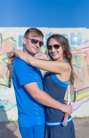 Two tennis players talking outdoors. Young man and young woman in tennis sportswear with tennis racquets in hands hugging looking at cameraの写真素材