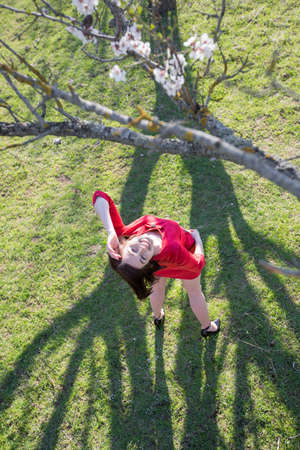 Girl in a neglected almond orchard. Attractive young woman in red dress standing with hand raised under blossoming almond tree and looking up smilingの写真素材