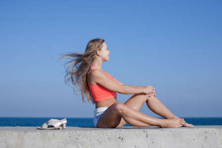 Girl at the sea. Barefoot young woman in pink short tank top with loose hair sitting on seafront looking awayの写真素材