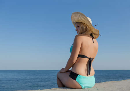 Girl at the sea. Young woman in swimsuit and straw hat sitting on concrete seafront looking awayの写真素材