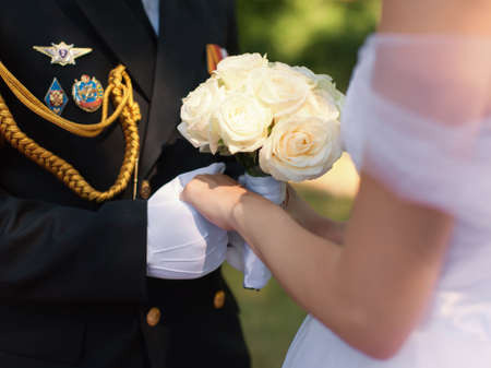 Wedding hands. Hands of the officer in white gloves and hands of his bride with a wedding bouquet from white roses during the wedding ceremony in the fresh airの写真素材