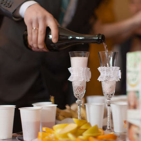 Man pouring champagne by the glasses. Glasses of the bride and groom, sliced fruit, champagne, juice, etc. on the tableの写真素材