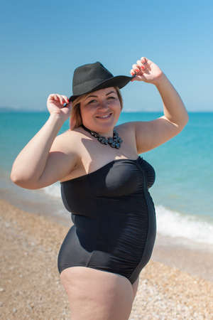 Overweight woman in black one-piece swimsuit and black hat stands against the beach with arms raised and looks at camera smiling. Portrait of fat girl in black swimwear at the seaの写真素材
