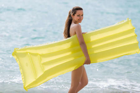 Young woman with yellow pool raft walks along sea. Smiling female person hides her nudity behind ...