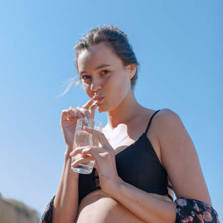 Girl in bikini against sky. Attractive young woman drinking water  from glass through a strawの写真素材