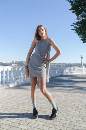 Portrait of girl on waterfront. Attractive female person in gray short sleeveless dress posing with feet inwards on paving slabの写真素材