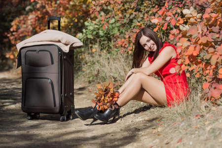 Young woman in red short dress resting in fall forest. Female person with bouquet from autumn leafs sitting on side of country road against colorful bushesの写真素材