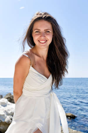 Portrait of funny long-haired girl in wedding dress against seashore. Dark-haired young woman in white sleeveless dress posing hiding hands behind back. She looks at camera and smiles in the backlightingの写真素材