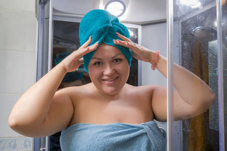 Portrait of girl in bathroom. Overweight woman wrapped in a towel with a turban on her head poses in a shower stall with her hands raisedの写真素材