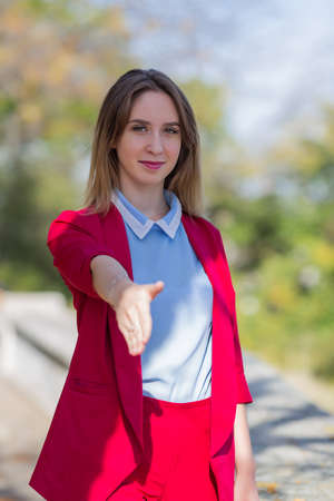 Portrait of blond woman in red suit outdoors. Young businesswoman holds out her hand for a handshake in autumn parkの写真素材