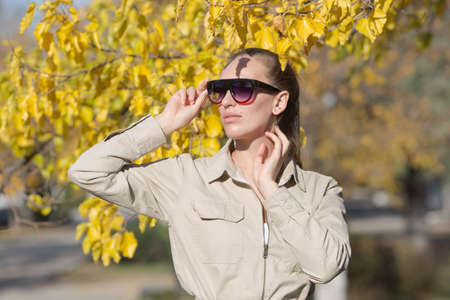 Waist up portrait of blonde person in sunglasses outdoors. Attractive young woman in tinted sunglasses and overalls similar to the uniform posing under autumn tree with hands raisedの写真素材