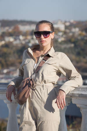 Three quarter length portrait of girl with shoulder bag in a jumpsuit outdoors. Attractive young woman in tinted sunglasses and overalls posing leaning on balustrade and looking at cameraの写真素材
