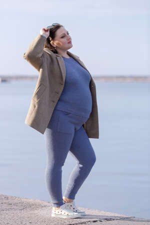Expectant mother on seafront. Pregnant woman in sportswear and cloth jacket posing against seaの写真素材