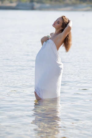 Expectant mother bathes on wild beach. Pregnant woman in white sarong poses knee-deep in waterの写真素材