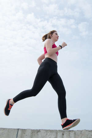 Active girl in sportswear running outdoors. Young sportswoman in black leggings and red top jogging...