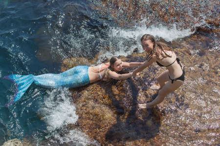 Two slim young women playing in shallow water. Girl in a black bikini pulls a friend in a mermaid costume out of the waterの写真素材