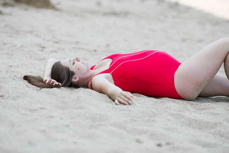 Pregnant woman in red one-piece swimsuit resting on sand beach in early time. Expectant mother lies on back sand in shadow of shoreの写真素材