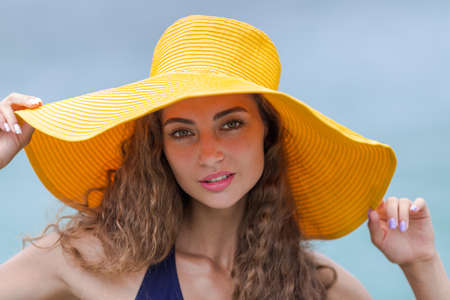 Portrait of pretty girl against sea. Curly young woman in yellow board brim hat poses with arms raised and looks at camera seriousの写真素材