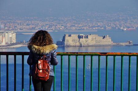 Aerial view of Naples and its gulf, with the Vesuvius in the background. A young woman standing on a balcony and watching the scenery. Winter clothes.のeditorial素材