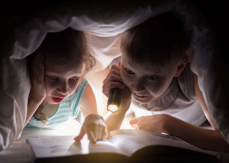 Children bedtime. Sister and brother are reading a book under a blanket with flashlight. Pretty young boy and lovely girl having fun in children room.の写真素材