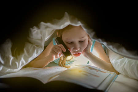 Children bedtime. Sister are reading a book under a blanket with flashlight. Pretty young girl having fun in children room.の写真素材