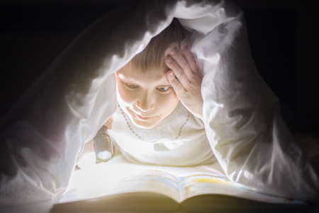 Children bedtime. Sister and brother are reading a book under a blanket with flashlight. Pretty young boy and lovely girl having fun in children room.の写真素材