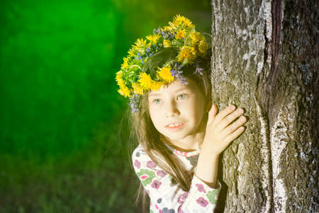 Cute girl in flowers wreath is looking out of birch tree in forest. The young witch.の写真素材