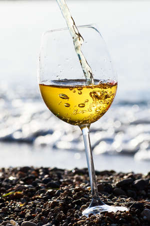 Alcoholic beverage of white wine pouring into glass outdoor on summer day on blue sea.