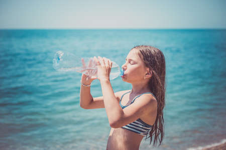 Cute little girl drinks water from bottle near the sea. Healthcare, lifestyle concept.の写真素材