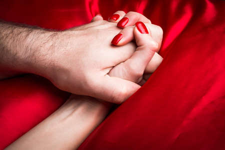 Young couple holding hands sensually on red silk bed.