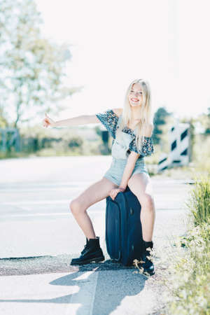 Beautiful young blonde girl smiling on the sunny road. Sitting on a suitcase luggage . Traveling the world with hitchhiking.の写真素材