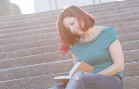 Handsome redhead young woman sitting on the stone stairs and  reading a book. - Concept Relaxing in nature.の写真素材