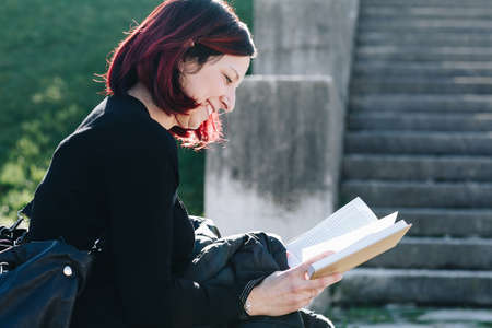 Handsome dark redhead young woman sitting on a small wall and reading a book in the public garden - Concept Relaxing in nature.の写真素材
