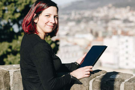 Happy smilling handsome redhead young woman sitting on a small wall working on tablet - Concept...