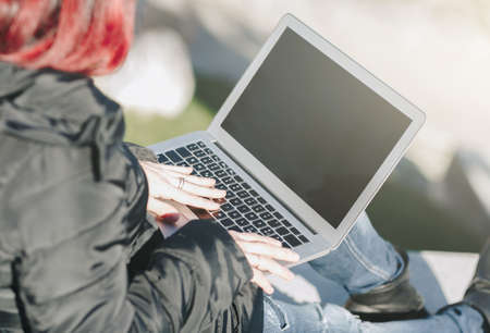 Happy smilling handsome redhead young woman sitting on a small wall and working on notebook -...