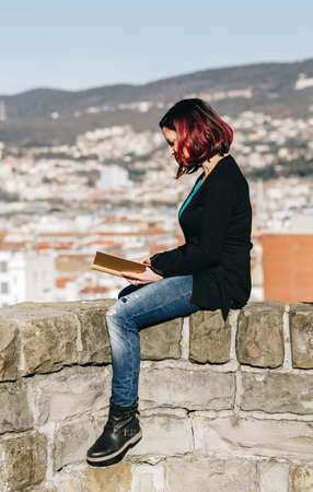 Handsome redhead young woman sitting on a small wall and reading a book with city in the background - Concept Relaxing in nature.の写真素材