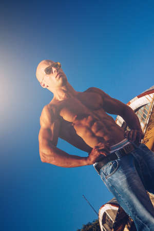 Beautiful male with blue eyes and sexy muscular body wear denim jeans and gold sunglasses with torso. Fitness male with healthy tan posing.の写真素材