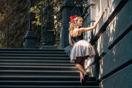 Elegantly dressed figure in polka dot skirt and black corset with red bow hair accessory poses dramatically on stone staircase, creating nostalgic 1950s aesthetic in urban setting.の素材