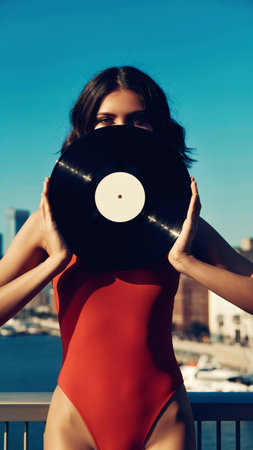 Stylish woman in vibrant red swimwear holds vintage vinyl record covering face against turquoise sky and city skyline. Perfect for music, fashion, lifestyle campaigns.の素材